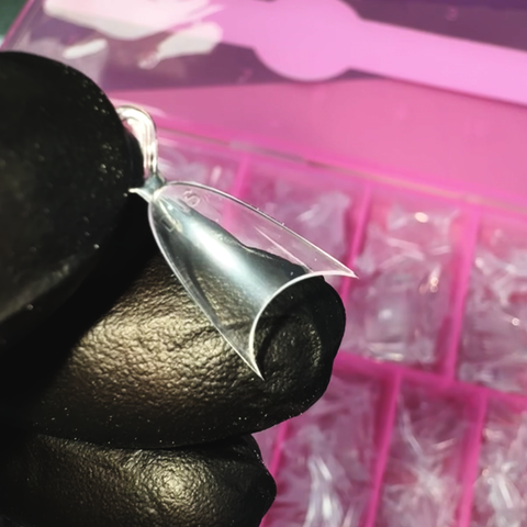 A close-up of a hand in a black glove holds one of Dynamic Nail Supply's Short Almond Nail Tips - Half Cover, highlighting its elegant curve. The background displays a pink organizer box filled with these flexible material nail tips, all translucent enough to reveal the gloved fingers beneath their clear surface.