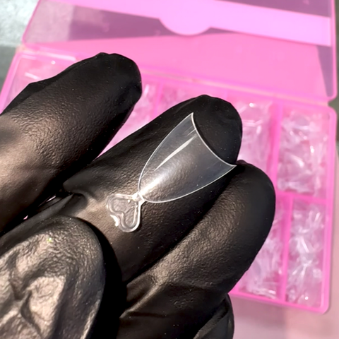 A hand in a black glove holds a transparent, hourglass-shaped nail stencil made from flexible material. In the background, a vibrant pink storage box with multiple compartments from Dynamic Nail Supply contains similar stencils and Short Almond Nail Tips - Half Cover (600 pcs in 12 sizes), also known as Short Stiletto tips. The contrast between the black gloves and the delicate plastic of the stencil and bright box is striking.