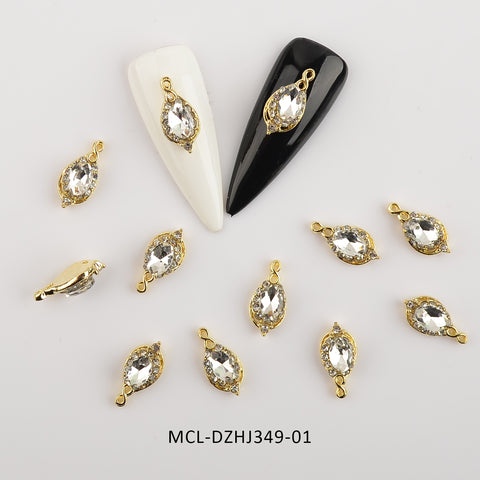Twenty pieces of unique shape charms for nail art design from Dynamic Nail Supply are scattered on a white surface. Two display nails, one white and one black, each showcase a charm featuring a crystalline center with a gold border and loops for attachment. The code MCL-DZHJ349-01 is visible.