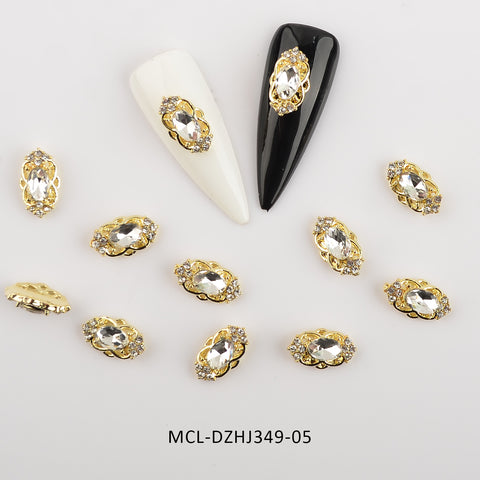 A dazzling collection of Dynamic Nail Supply's "20 pcs / 1 bag of Unique shape Charms for Nails Art Design" featuring oval-shaped gold and rhinestone decorations is artfully spread across a white background. Two false nails, one white and one black, each showcase a rhinestone charm from the collection. Displayed prominently are these exquisite designs with the code MCL-DZHJ349-05 printed below them.
