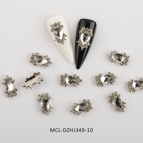 A close-up of decorative nail art pieces highlights shimmering, jewel-adorned embellishments. Two artificial nails, one in white and the other in black, display unique shape charms from Dynamic Nail Supply's "20 pcs / 1 bag of Unique shape Charms for Nails Art Design." Several loose pieces are scattered around the nails. Product code MCL-DZHJ349-10 is shown at the bottom.