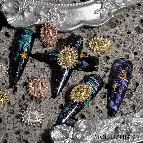 A set of long, black, coffin-shaped nails is showcased on a textured gray surface. The nails exhibit vibrant blue and green marbling highlighted by gold stars and crescents, adorned with 12-piece Virgin Mary charms for religious nail designs from Dynamic Nail Supply. Decorative gold and silver accents enhance the display, with an ornate silver frame positioned nearby to complement the elegant and intricate style.