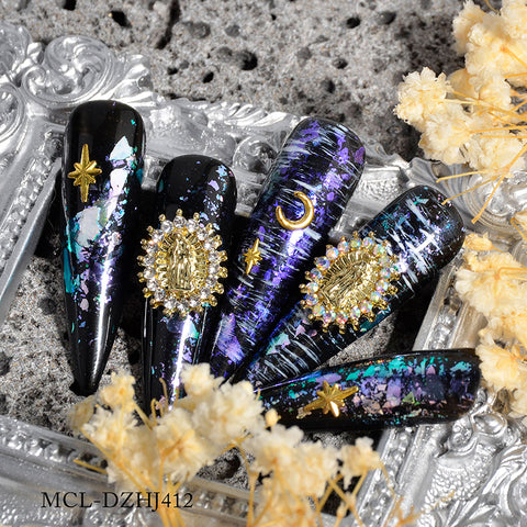 A close-up showcases intricate nail art on long, black stiletto nails, adorned with iridescent purple and blue foil accents. The designs include gold celestial shapes and ornate embellishments such as stars, complemented by a Virgin Mary charm from the (Small version) 12 pcs bag by Dynamic Nail Supply. A detailed gold figure encircled by pearls and nearby dried flowers complete the look.