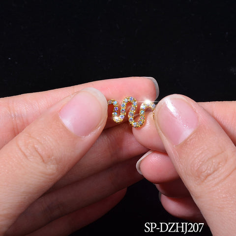 A close-up image features hands holding a small, gold, snake-shaped charm encrusted with colorful gemstones against a black background. The nails are neatly trimmed and clean, suggesting an exquisite nail art design. In the bottom right corner, white text reads "Dynamic Nail Supply - 10 pcs of Snake Shape Charms for Nails Art Design (Smaller version).