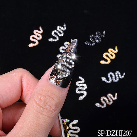 A hand showcases a metallic silver nail art design featuring a dazzling, rhinestone-studded snake, complemented by snake-shaped charms in vibrant metallic tones on a black surface. A lens flare accentuates the sparkle of the rhinestones. The product is identified as the 10-piece set of Snake Shape Charms for Nail Art Design (Smaller version) by Dynamic Nail Supply.