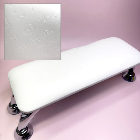 A white, rectangular full-grain leather armrest cushion pillow by Dynamic Nail Supply, featuring a subtle embossed LV pattern reminiscent of elegant designs. It is supported by four shiny chrome legs and set against a pink background. An inset highlights the stylized floral and geometric motif, enhancing the charm of any manicure service setting.