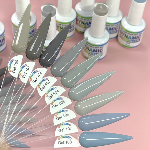 A set of six long, pointed nail samples in diverse shades of gray and blue, labeled Gel 103 to Gel 108, displays the (New) Fall Color Gel Polish Collection from Dynamic Nail Supply, arranged like a fan on clear sticks. Ideal for the Fall Season, they sit on a pink surface with blurred bottles in the background promising long-lasting wear.