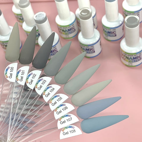 A set of pointed nail polish samples, featuring the (New) Fall Color Gel Polish Collection from 100 to 108 by Dynamic Nail Supply, is arranged in a fan shape on a pink surface. The polish colors range from light to dark gray and greenish-gray, ideal for the fall season. In the background are bottles of gel polish from Dynamic Nail Supply.