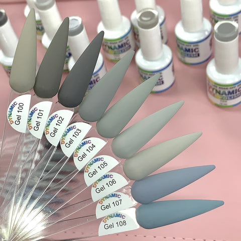 The image showcases nine elongated, almond-shaped nail samples in various shades of gray, labeled from (New) Fall Color Gel Polish Collection 100 to 108. These samples are elegantly displayed on a transparent holder. In the background, white bottles featuring Dynamic Nail Supply's vivid labels hint at long-lasting wear and are perfect for the Fall Season.