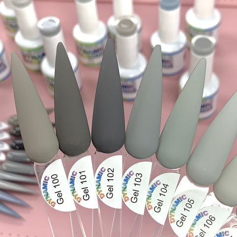 Close-up of seven samples from the Dynamic Nail Supply's (New) Fall Color Gel Polish Collection, labeled from 100 to 106, showcasing an array of gray shades. Each sample stick features a pointed end that ensures long-lasting wear. In the background, blurred bottles of nail polish rest on a pink surface, capturing the essence of fall colors.