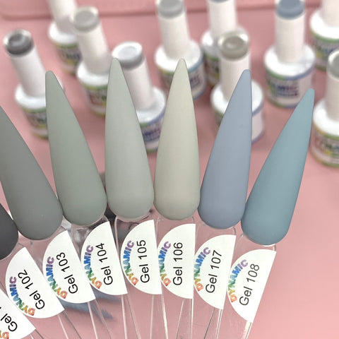 A close-up of a display featuring sample nail colors on pointed tips, labeled Gel 102 to Gel 108, showcases shades from the (New) Fall Color Gel Polish Collection by Dynamic Nail Supply. These colors range from dark gray to light grays and include a soft blue, making them perfect for the fall season. In the background, blurred bottles of polish rest on a pink surface.