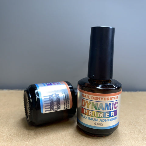 A compact brown bottle with a glossy black cap, labeled Dynamic Primer Nail Dehydrator Maximum Adhesion 13 mL, stands upright on a cardboard surface beside the Acrylic Nail Starter Kit for professional or beginner by Dynamic Nail Supply. Close by, another similar bottle is laying on its side, partially showing colorful text and designs that complement the powders inside.