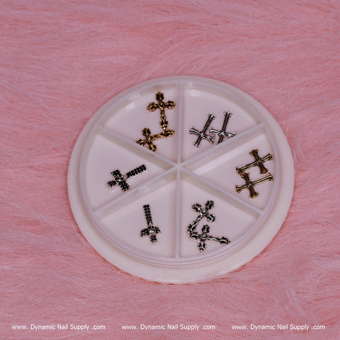 A circular container featuring six compartments on a pink textured background holds small, decorative 3D metallic cross-shaped charms in silver and gold, ideal for Halloween nail art. The base is adorned with the repeated print of www.DynamicNailSupply.com.