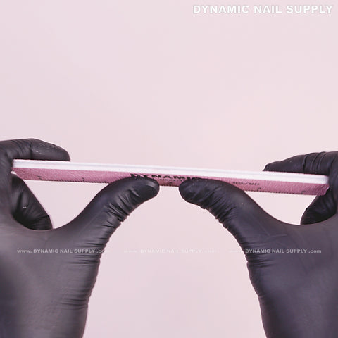 Gloved hands hold a Square Nail file 80/80 Grit, Pink, Double-sided, pack of 10 by Dynamic Nail Supply on pink background.