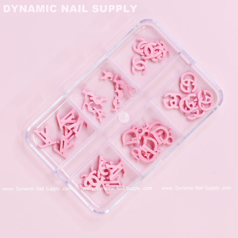 A clear plastic organizer showcases 30 pieces of pink multi-shaped resin brand name charms from Dynamic Nail Supply against a pastel pink background. Five compartments feature a variety of decorative pieces in whimsical shapes, with the company name prominently printed in white at both the top and bottom.