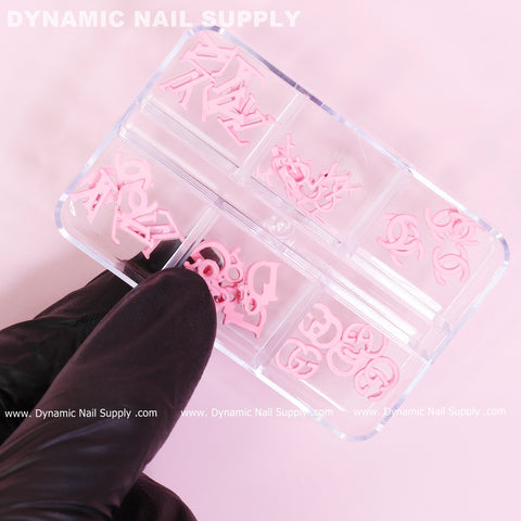 A hand in a black glove holds a clear plastic container with several compartments, each filled with 30 pieces of pink multi-shape resin brand name charms designed for nail art, featuring intricate designs such as swirls and abstract shapes. The words Dynamic Nail Supply elegantly stand out in the background.