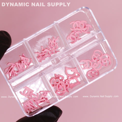 A hand wearing a black glove holds a transparent plastic box featuring six compartments filled with various pink nail art decorations, including enchanting 30 pcs of Pink Multi-shapes Resin Brand Name Charms for Nail Art Design. These compartments display abstract shapes and letters against a soft pink background. The box is from Dynamic Nail Supply.