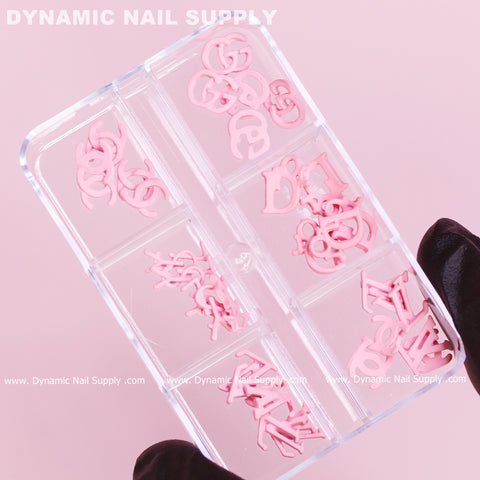 A gloved hand holds a clear plastic case containing eight compartments filled with the "30 pcs Pink Multi-shapes Resin Brand Name Charms for Nails Art Design," designed in letter shapes. The soft pink background enhances the elegance of the image, with "Dynamic Nail Supply" prominently displayed across the top and bottom edges, highlighting the brand's design prowess.
