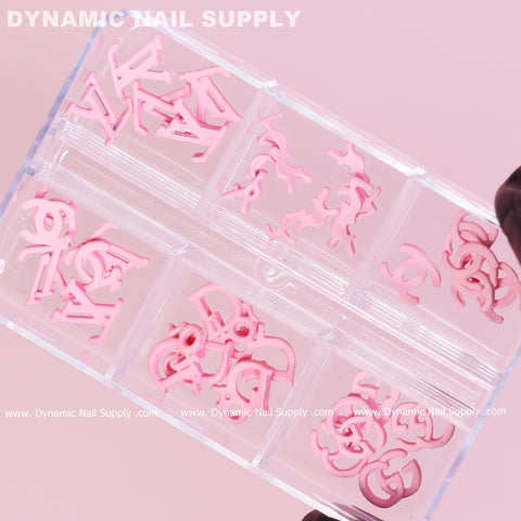 A hand with dark skin elegantly holds a clear plastic box containing 30 small, pink multi-shaped resin brand name charms for nail art design. The bottom of the box features the repeated logo "Dynamic Nail Supply.