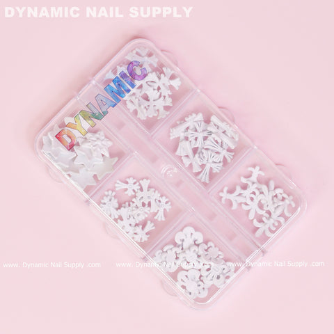 A clear plastic organizer on a pink background showcases white 3D nail art charms, including flowers, bows, and 60 pieces of matte white chrome heart charms featuring antique crosses. The lid features the Dynamic Nail Supply logo in colorful letters, adding flair to your nail art design collection.