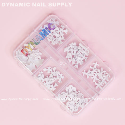 A clear plastic organizer contains a variety of white nail art decorations, including stars, bows, and matte white chrome heart charms shaped like antique crosses. The lid prominently features the word DYNAMIC in colorful letters. Encasing a soft pink gradient background, the name Dynamic Nail Supply is repeated along the bottom edge.