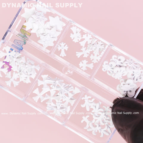 A person wearing a black glove is holding a clear container filled with matte white chrome heart charms for nail art design, shaped like stars, moons, flowers, and butterflies against a pink background. The container is neatly divided into compartments and features the Dynamic Nail Supply brand name.