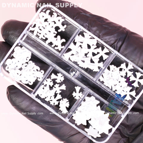 A hand in a black glove holds a transparent plastic container with eight compartments, featuring the 60 pcs Matte White Chrome Heart Charms (Antique Crosses) for Nails Art design. Inside, small, white decorative pieces shaped like bows and flowers complement the charms. This container, perfect for any nail art design enthusiast, is from Dynamic Nail Supply.