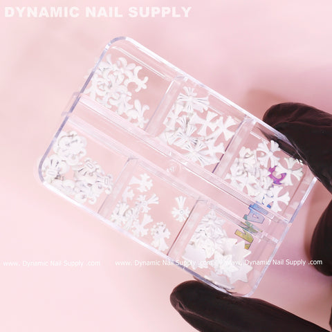 A person wearing black gloves holds a transparent box with small compartments filled with various white nail art decorations in floral shapes. Among them, the 60 pcs Matte White Chrome Heart Charms (Antique Crosses) for nail art design stand out. The soft pink background highlights the brand name Dynamic Nail Supply along the edges.