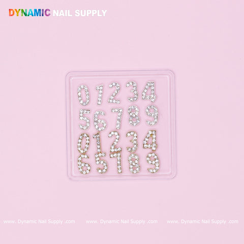 A square container on a pink background features rhinestone-studded digits from 0 to 9, organized in four rows. Each charm from the "20 pcs Gold and Silver 3D Number Charm with Rhinestone (0 to 9)" set gleams with a silver-like finish against a transparent base. The brand name "Dynamic Nail Supply" is displayed in colorful letters at the top left corner.