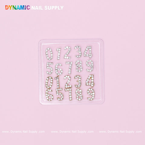 A clear plastic case contains the "20 pcs Gold and Silver 3D Number Charm with Rhinestone (0 to 9)" from Dynamic Nail Supply, featuring dazzling rhinestone-covered metal numbers arranged in three neat rows on a pink background, complemented by elegant silver and gold 3D number charms. The brand name, Dynamic Nail Supply, is prominently displayed in vibrant letters at the top left.