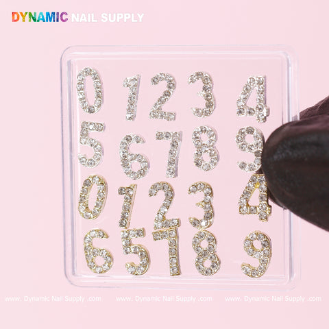 A gloved hand holds a clear plastic square from Dynamic Nail Supply against a soft pink background, displaying the 20-piece Gold and Silver 3D Number Charm with Rhinestones. The decorative numerals, ranging from 0 to 9 along with an extra 4 and 5, are arranged in two rows.