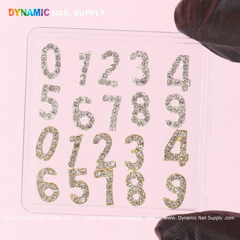 A transparent square sheet showcases two sets of numbers 0-9, each embellished with petite, shimmering rhinestones and accented by elegant Gold 3D Number Charms. "Dynamic Nail Supply" is prominently printed in vibrant text at the top-left corner. The sheet is held up by black-gloved fingers against a soft pink backdrop.