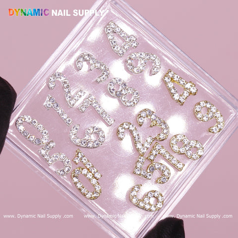 A clear container beautifully displays the 20-piece set of Gold and Silver 3D Number Charms with Rhinestone, featuring numbers 0 through 9 outlined in gold against a pink background. A black-gloved hand holds this sparkling collection, while the brand name "Dynamic Nail Supply" is prominently featured at the top in multicolored letters.