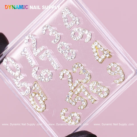 A clear plastic case showcases sparkling, rhinestone-studded numbers from 0 to 9. Each number in the Gold and Silver 3D Number Charm with Rhinestone set features a gold outline and is arranged on a soft pink background. The brand name, Dynamic Nail Supply, is partially visible at the top in colorful letters.