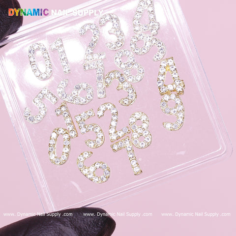 A gloved hand holds a clear plastic container displaying the 20 pcs Gold and Silver 3D Number Charm with Rhinestone (0 to 9) by Dynamic Nail Supply. These sparkling rhinestone numbers, ranging from 0 to 9, are randomly arranged against a light pink background. The Dynamic Nail Supply logo is visible at the top left and bottom center.