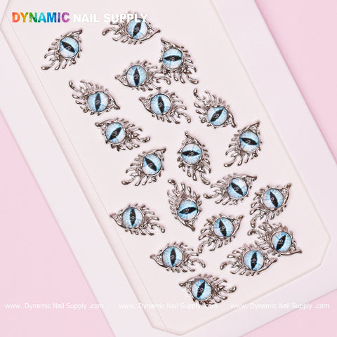 A set of The Light Blue GreaT EyEs (Eye of SauR0n) charms for nail art design, featuring metallic silver frames and blue centers, arranged on a white rectangular surface. The pastel pink background highlights the vibrant "Dynamic Nail Supply" lettering at the top.