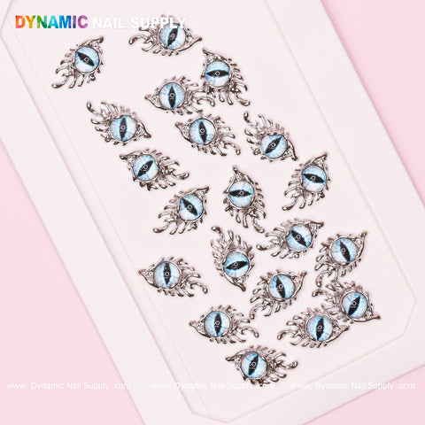 An assortment of The Light Blue GreaT EyEs charms for nail art design, featuring Eye of SauR0n motifs with silver frames, is displayed in rows on a white rectangular tray. These charms resemble eyes with intricate metal detailing resembling lashes. The name "Dynamic Nail Supply" is prominently displayed at the top, accompanied by repeated website URLs at the bottom.