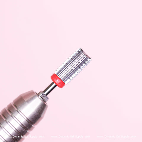 A close-up shot captures the Dynamic Nail Supply's new Safety Barrel Bit, featuring a fine grit for smoothing acrylics and sealing cuticles. The cylindrical, textured bit is distinguished by a red band near the top and is attached to a silver, ridged handle. Set against a soft pink background, it offers a clean and minimalist aesthetic.