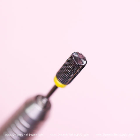 A close-up image features the Super Fine Barrel Bit from Dynamic Nail Supply, showcasing its silver cylindrical design with extra fine grit and a textured surface. The bit, attached to a drill handle, has a yellow band near the base, making it ideal for cuticle sealing. The soft pink background complements the company's watermark at the bottom edge.