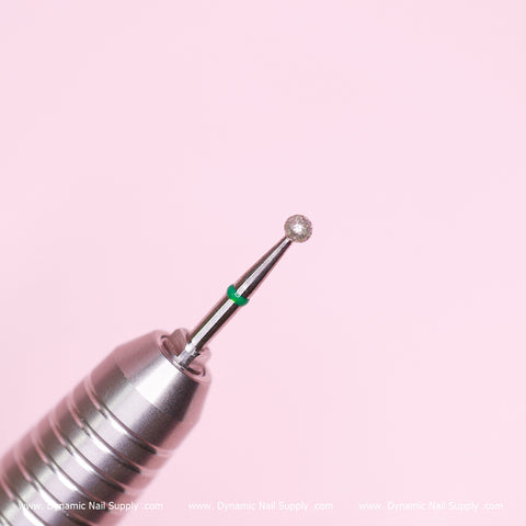 A close-up of the Coarse Cuticle Drill Bit by Dynamic Nail Supply, featuring a non-abrasive silver ball bit with a rounded tip and green band, set against a soft pink background. The grooved handle indicates its ideal use for cuticle cleaning and removing dead skin during nail art or manicure tasks.