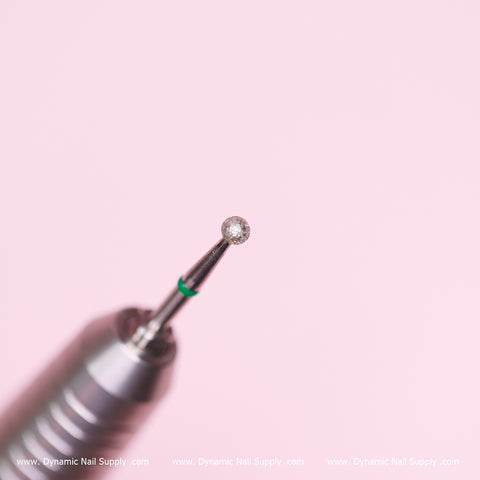 A close-up showcases a silver Coarse Cuticle Drill Bit from Dynamic Nail Supply, featuring a textured spherical tip with a 0.12-inch diameter against a soft pink backdrop. Ideal for cuticle cleaning and removing dead skin, the drill bit's shaft is adorned with a green band near the tip. The branding "www.DynamicNailSupply.com" is elegantly displayed at the bottom in white.