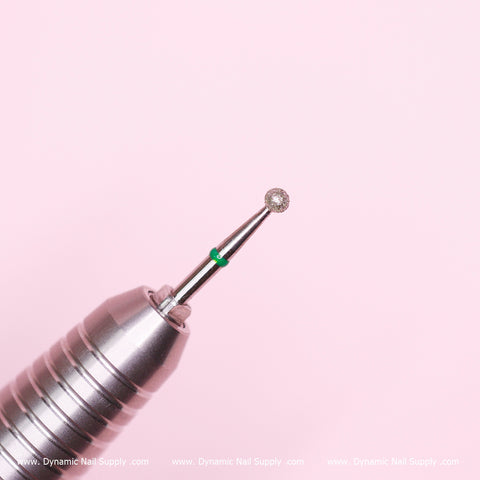 A close-up of a Coarse Cuticle Drill Bit by Dynamic Nail Supply, set against a gentle pink background, highlights its diamond-like spherical tip designed for gentle cuticle cleaning and removing dead skin. A green ring adorns the base, underscoring its precision in manicures.