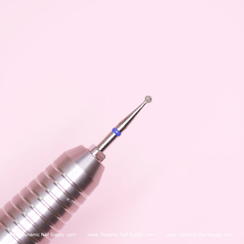 A close-up of a metallic Medium Cuticle Drill Bit from Dynamic Nail Supply showcases its textured, rounded tip, ideal for cuticle area cleaning, against a soft pink background. The tool features grooved sections and a blue marking near the tip. Partial text at the bottom edge reads: Dynamic Nail Supply.