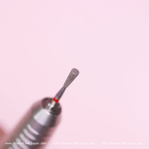 Close-up of a metallic nail drill bit against a soft pink background. This fine, textured cylindrical drill bit by Dynamic Nail Supply is designed for precision in nail care, making it perfect for prepping and cleaning the cuticle area as well as removing dead skin. The bottom edge features www.DynamicNailSupply.com.