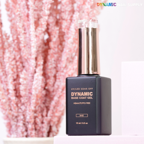 A black bottle of Dynamic No-HEMA Base Coat Gel with rose gold cap sits before pink plants for gentle, healthy bonding.