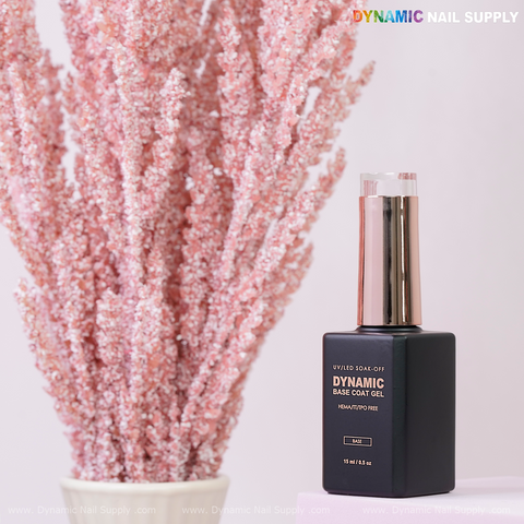 A bottle of Dynamic No-HEMA Base Coat Gel sits beside pink dried flowers on a light pink background.