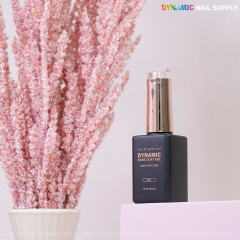 A bottle of Dynamic No-HEMA Base Coat Gel for healthier nails stands on a pink surface with flowers in the background.