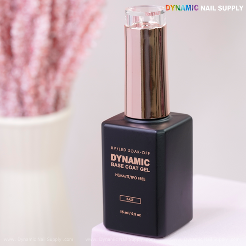 A black bottle of Dynamic No-HEMA Base Coat Gel with a rose gold cap, on a white surface, pink decor blurred behind.