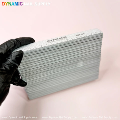 A gloved hand holds a stack of gray nail files against a soft pink background. The top file, perfect for shaping acrylic nails, showcases the brand Dynamic Nail Supply with grit numbers 80/100. These slightly curved rectangular files, known as Strong and Hard professional Jumbo nail files (25 pcs), have a smooth texture ideal for precise manicures.