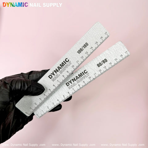 A gloved hand holds two gray files from the "Strong Emery Board - Professional Acrylic nail files" set by Dynamic Nail Supply, ideal for achieving flawless acrylic nails. Each file in the set of 25 features a grit of 100/180, and is labeled with "Dynamic Nail Supply" at both ends in assorted colors, all against a pale pink backdrop.